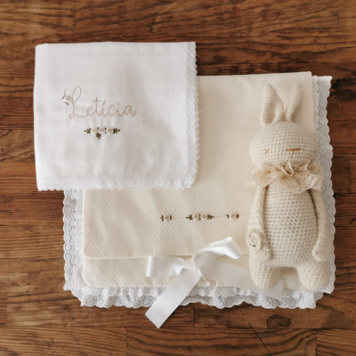Fralda branca bordada com nome Letícia, manta creme com renda branca e brinquedo peluche creme sobre madeira