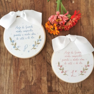 Dois aros de bordado com bordados florais e texto, com laços brancos, sobre madeira e flores coloridas ao lado
