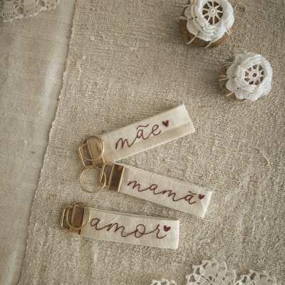 Três porta-chaves de tecido bege com palavras bordadas e dois frascos decorados com renda branca sobre tecido rústico