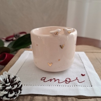 Vaso de cerâmica branca com corações dourados sobre tecido branco bordado com a palavra 'amor' e pinha pintada de branco na mesa de madeira
