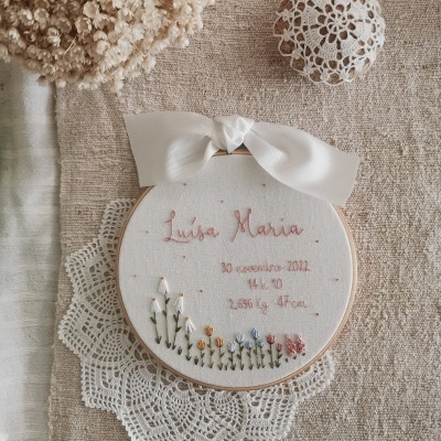 Aro de bordado com nome, data e medidas, flores bordadas e laço branco sobre fundo bege com renda branca