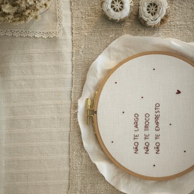 Bastidor de bordado com texto em tecido branco e botões decorativos em crochê sobre tecidos beges.