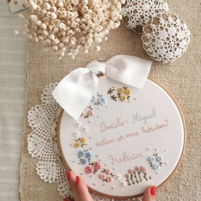 Bastidor de bordado decorado com flores e texto, com laço branco, sobre tecido bege com flores secas e bolas de croché