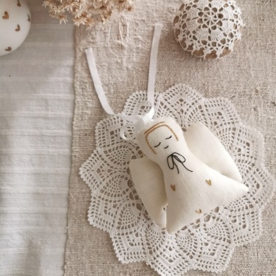 Boneco anjo de tecido branco com detalhes dourados e ribbon preto, sobre renda branca em superfície clara