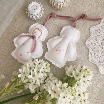 Dois anjos decorativos de tecido branco com fitas rosa, ovos decorativos em rede branca e flores brancas sobre superfície bege.