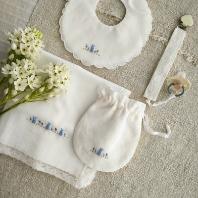 Conjunto de bebé branco com bordado azul e mola para chupeta sobre tecido bege com flores.