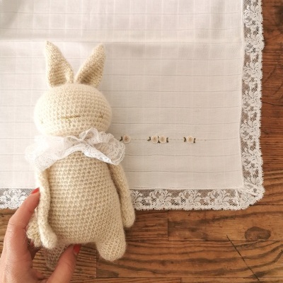 Boneco de coelho em crochê bege com laço de renda branca sobre pano branco com borda de renda numa mesa de madeira.