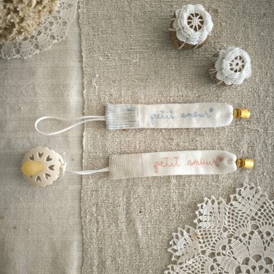 Pega-chupetas em tecido com texto 'petit amour' e frascos pequenos com cobertura em renda sobre tecido bege e rendas.