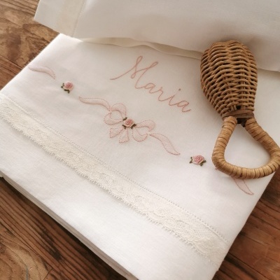 Toalha branca bordada com o nome Maria, rosas e laço, com detalhe de renda, ao lado de maraca de palha
