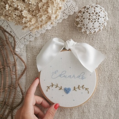 Bastidor de madeira com bordado do nome Eduardo, coração azul e laço branco sobre mesa de linho