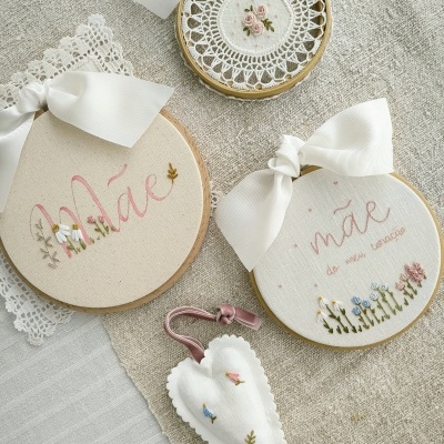 Peças decorativas bordadas à mão em tecido bege e branco com laços e flores, incluindo bastidores com a palavra 