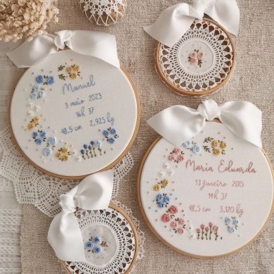 Quadros decorativos em bastidores de madeira com bordados florais e informações pessoais, com laços brancos, sobre toalha de linho.