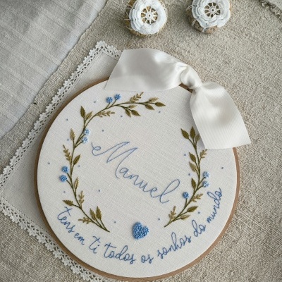 Aro de bordado com texto 'Manuel' bordado e decoração de flores azuis com laço branco.
