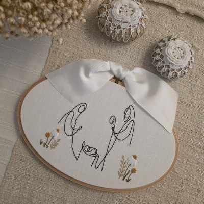 Bordado minimalista de figuras humanas com bebê em bastidor com laço branco.