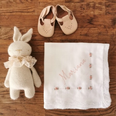 Brinquedo de crochê de coelho branco, sapatos de pele bege para bebé e pano branco com bordado cor-de-rosa num fundo de madeira