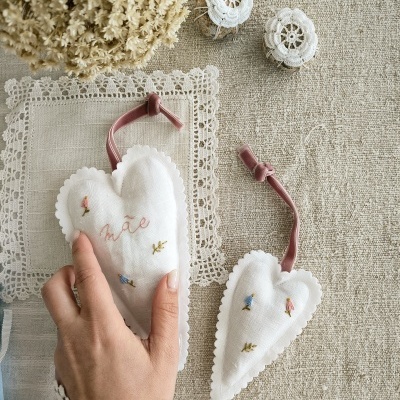 Dois corações brancos de tecido com bordados coloridos e fitas de veludo rosa, sobre tecido cru e renda, com flores secas ao lado.