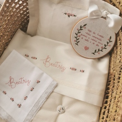 Conjunto de roupa de cama branca bordada com nome Beatriz e motivos florais numa cesta de verga.
