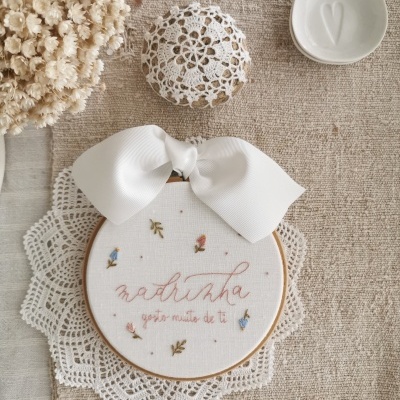 Quadro bordado com texto para madrinha, flores pequenas, laço branco, sobre tecido bege e renda branca com flores secas ao lado