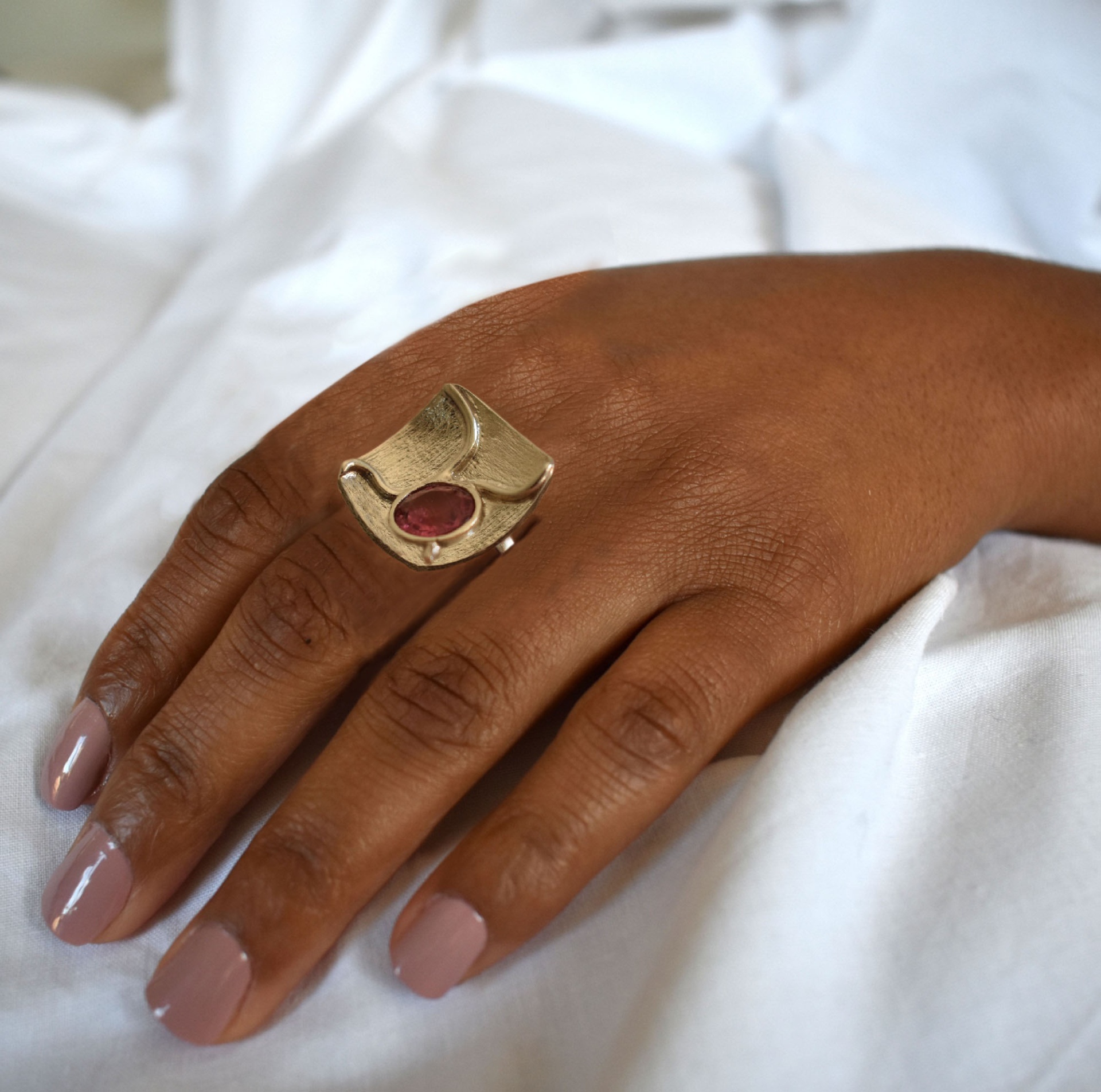 Anel June Anel grande dourado com pedra vermelha numa mão com unhas rosa claro sobre tecido branco