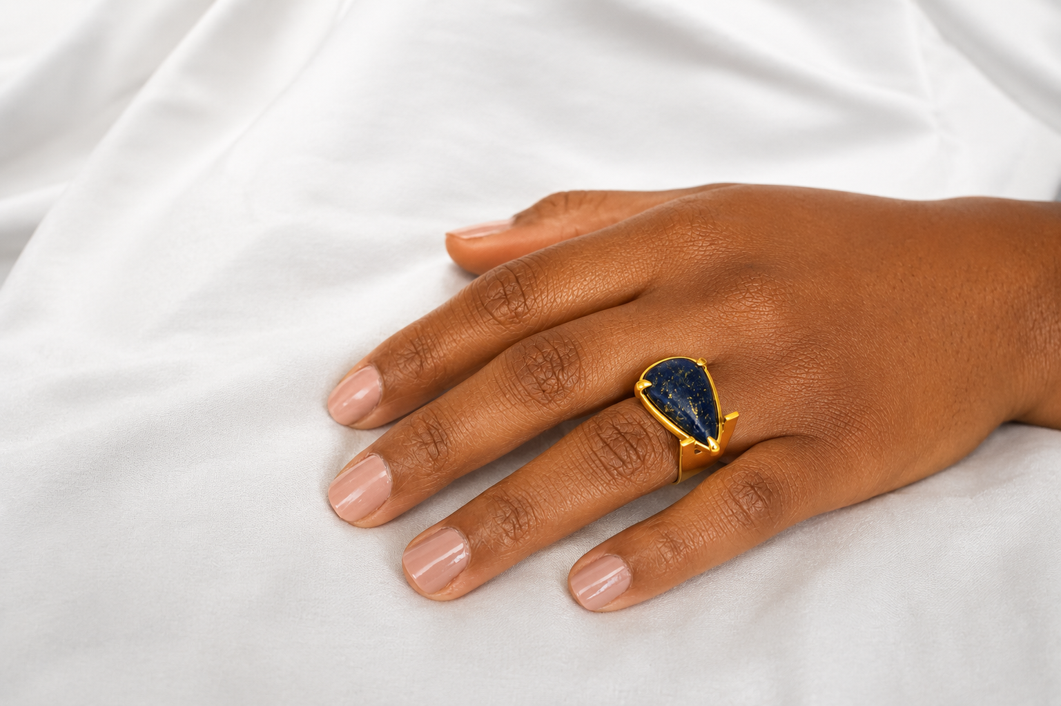 Anel assimétrico Anel dourado com pedra azul escura em forma de gota na mão sobre tecido branco