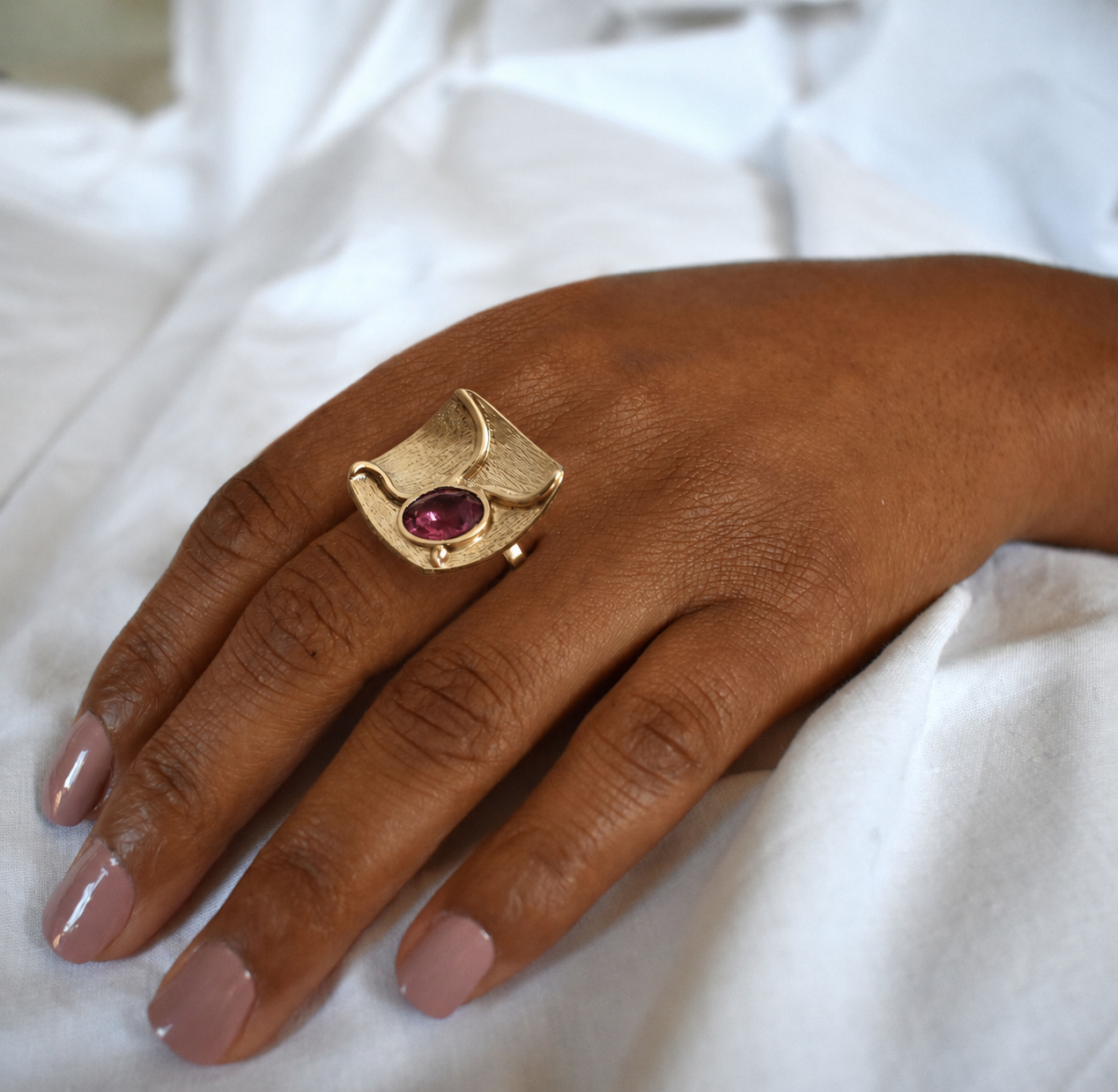 Anel June Mão com anel dourado com pedra roxa sobre tecido branco