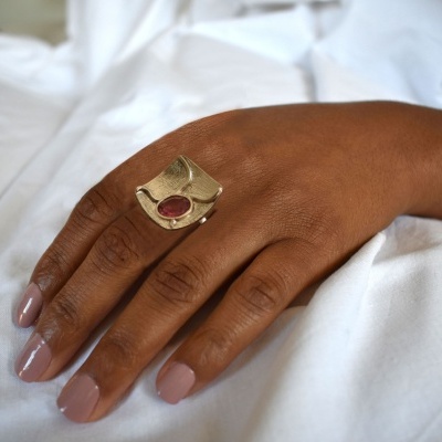 Anel grande dourado com pedra vermelha numa mão com unhas rosa claro sobre tecido branco