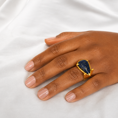 Anel dourado com pedra azul escura em forma de gota na mão sobre tecido branco