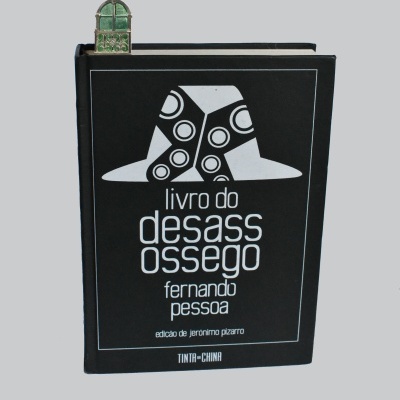 Capa preta de livro 'livro do desassossego' com ilustração de chapéu, marcador metálico verde em forma de janela