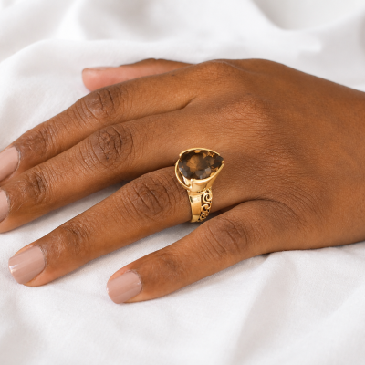 Anel dourado com pedra castanha em forma de gota na mão feminina sobre tecido branco