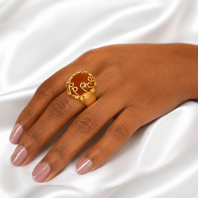 Anel dourado com pedra âmbar numa mão feminina sobre tecido branco.