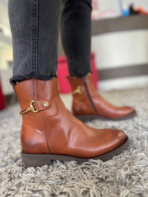 Botim ABF-97 Pisa230 Botas castanhas em pele com fivela dourada usadas com calças pretas sobre tapete cinza