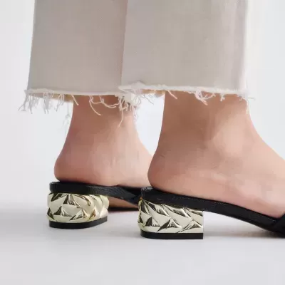 Sandálias pretas com salto dourado trançado usadas com calças bege claras desfiadas