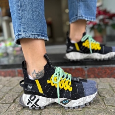 Ténis desportivo preto com detalhes amarelos e azul claro, atacadores verdes, usado por pessoa com jeans azuis