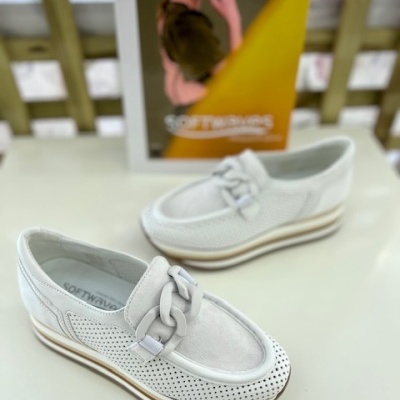 Sapatos mocassim brancos com lacinho e perfurações, em superfície branca com cartaz ao fundo