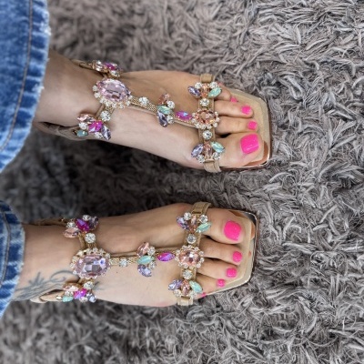 Sandálias bege com pedras coloridas em pés femininos com unhas pintadas de rosa fluorescente sobre tapete cinzento felpudo.