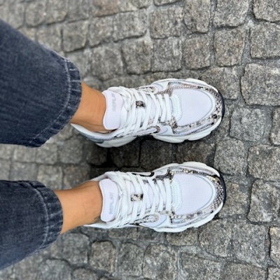 Ténis brancos com padrão pele de cobra usados com calças pretas num chão de pedras