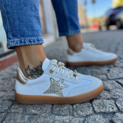 Ténis brancos com detalhes dourados e estrela prateada, usados com calças de ganga