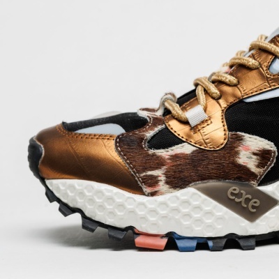 Tênis de corrida com couro bronze metálico, pelo castanho e branco, sola branca texturada e detalhes coloridos
