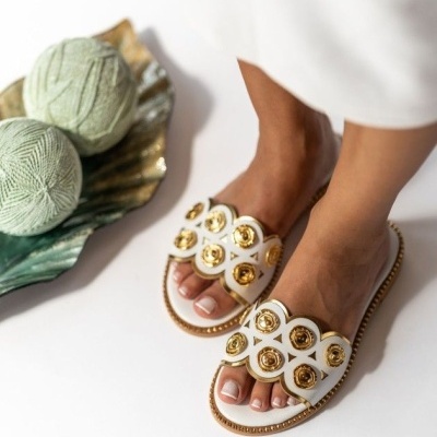Sandálias brancas com detalhes dourados usadas por uma pessoa em fundo branco com prato decorativo