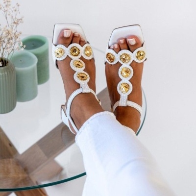 Sandálias brancas com detalhes dourados em tecido de textura, pernas com calças brancas junto a uma mesa de vidro e velas verdes.