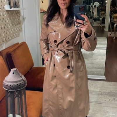 Mulher com casaco bege brilhante de comprimento médio, cinto e botões pretos, tirando selfie num espelho numa sala elegante.