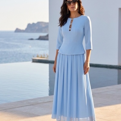 Mulher com vestido azul claro comprido e mangas três quartos junto a uma piscina e mar ao fundo