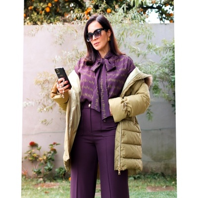 Mulher vestida com casaco acolchoado bege, blusa roxa com laço e calças largas roxas, a usar óculos de sol e segurando um telemóvel