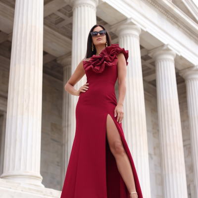 Mulher com vestido vermelho longo com folhos num ombro em frente a colunas de mármore branco