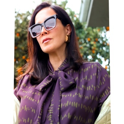 Mulher com blusa roxa com padrão verde e laço, óculos de sol grandes pretos e brincos dourados