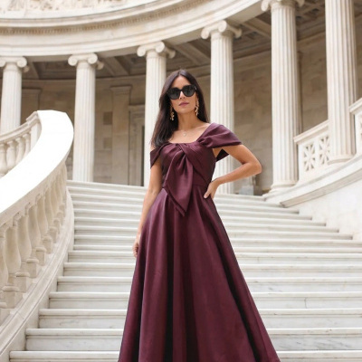 Vestido comprido vinho com nó frontal, sandálias douradas, óculos escuros, escadaria de mármore e colunas clássicas.