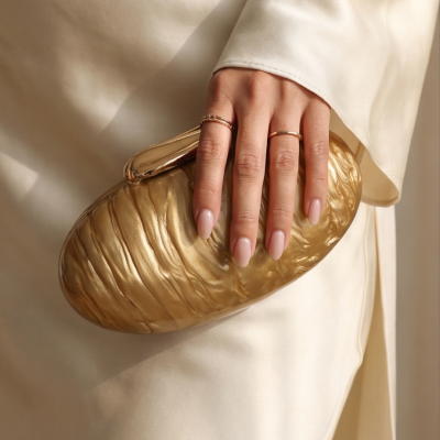 Mão segurando mala dourada ondulada com anéis dourados e roupa creme acetinada