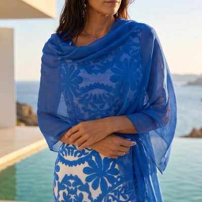 Mulher com vestido branco e azul e estola azul translúcida perto de piscina com mar ao fundo.