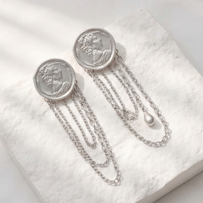 Brincos prateados com medalhões e correntes pendentes sobre superfície branca