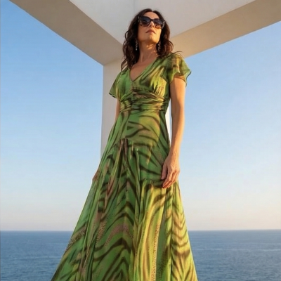 Mulher com vestido verde comprido com padrão, óculos escuros, ao ar livre com vista para o mar